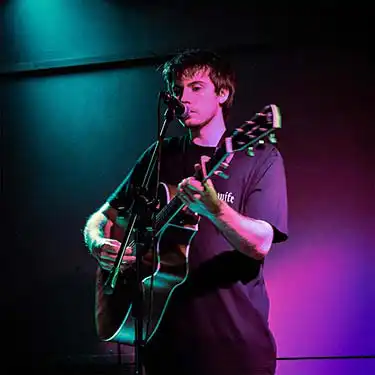 Image of the artist with guitar performing in an indoor venue.